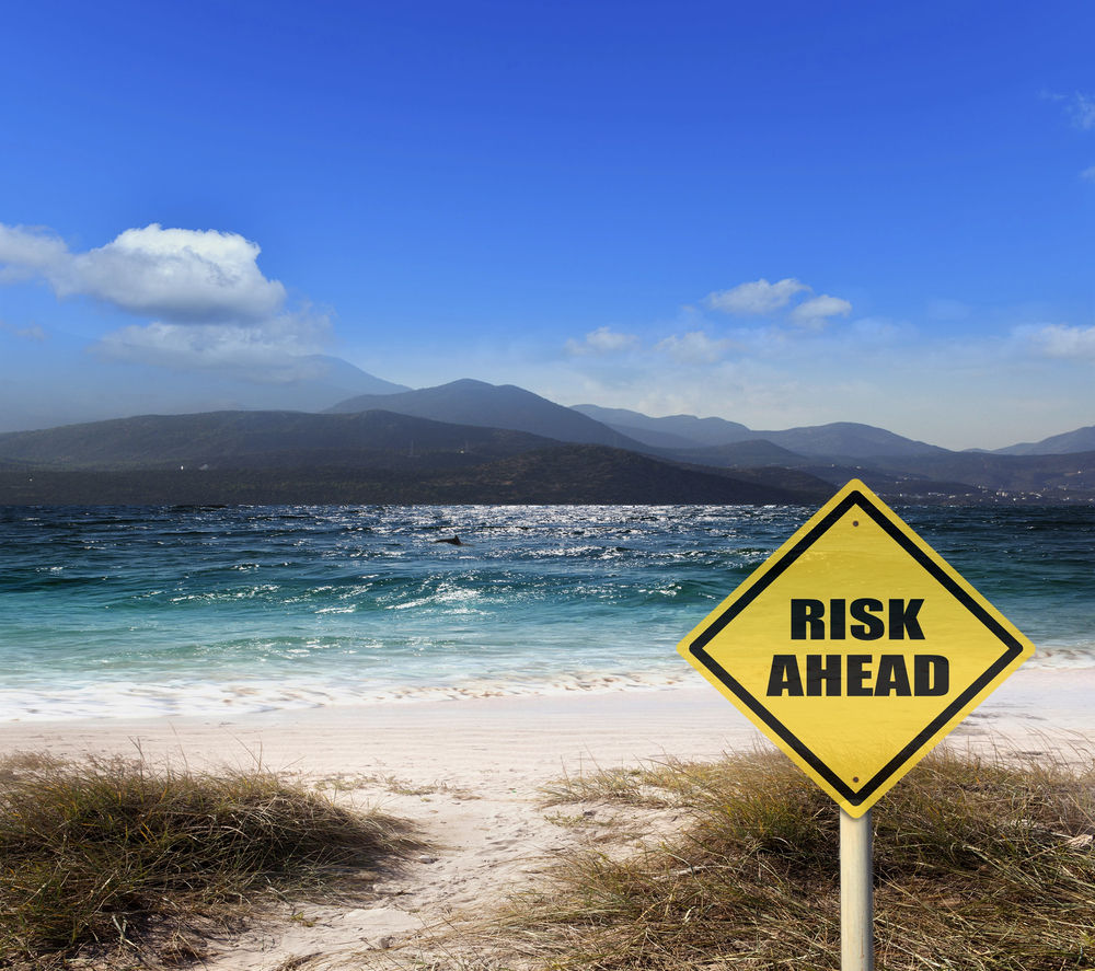 A yellow diamond-shaped sign reading RISK AHEAD stands on a grassy beach, facing the sea, with mountains and a partly cloudy blue sky in the background.