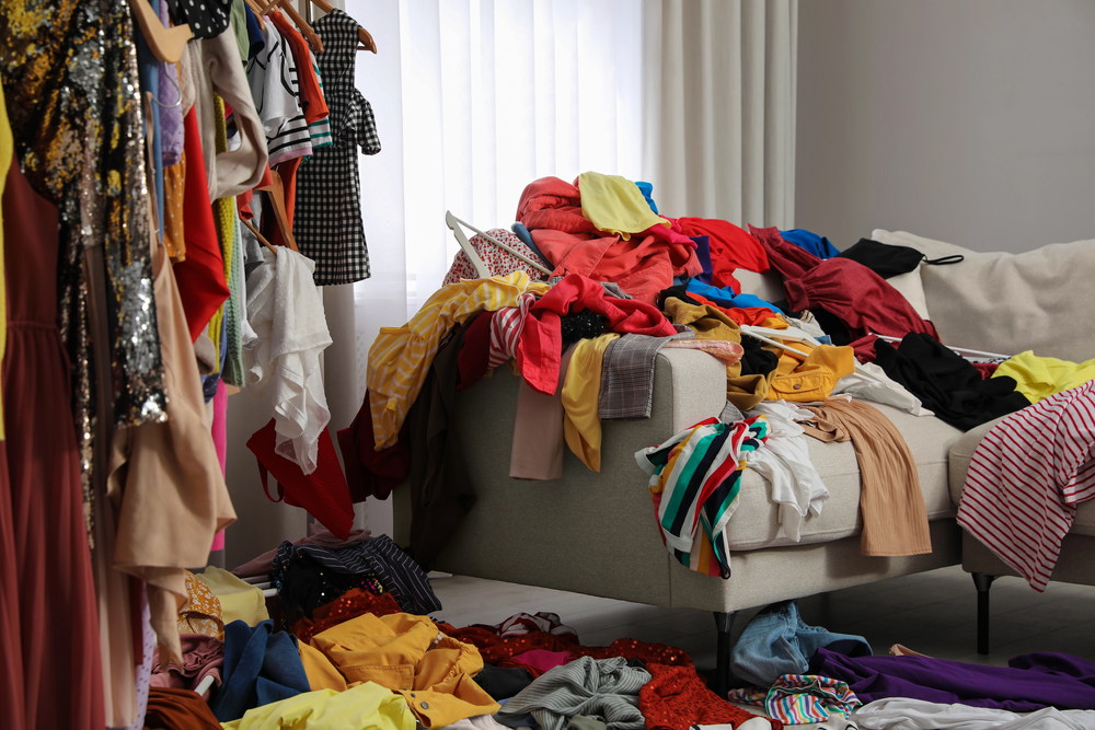 A messy room with colourful clothes piled on a beige sofa, scattered on the floor, and hanging on a clothes rail near a window with sheer curtains.