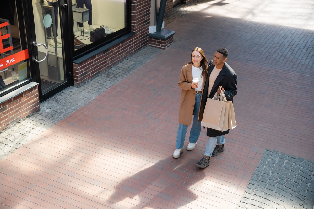 A young couple walk together on a brick path, holding shopping bags and a coffee cup. They are both wearing coats and smiling, passing by a shop with a red sale sign in the window.