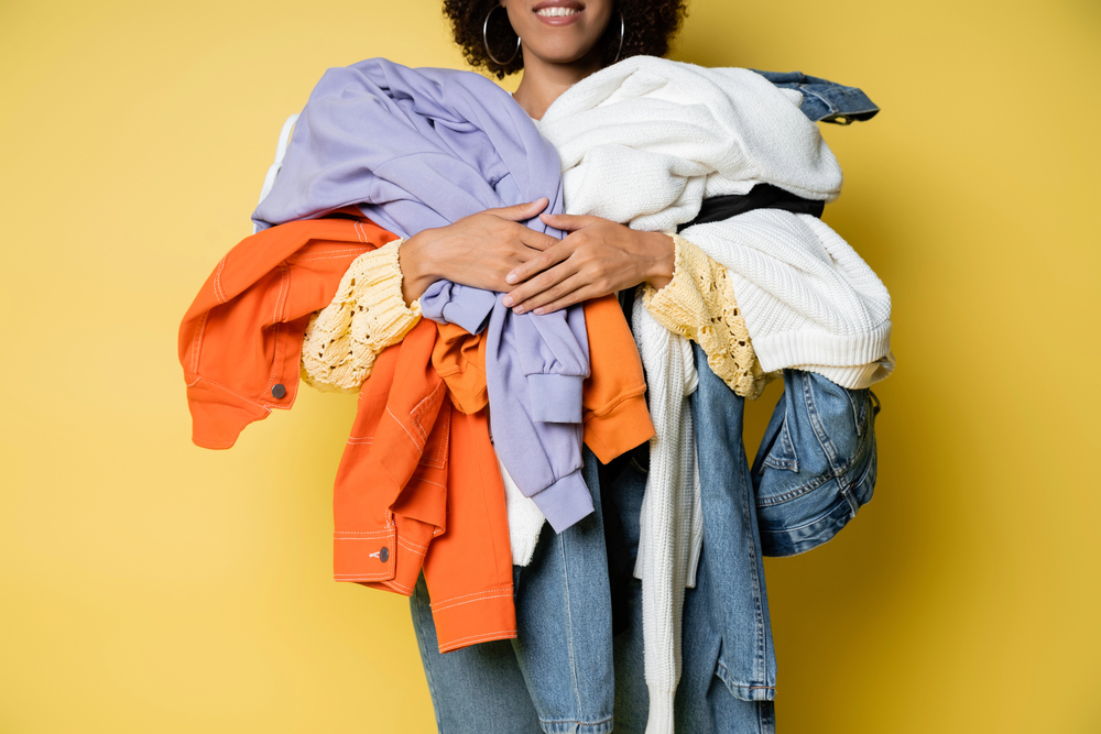 A person holding a large pile of colourful clothes, including orange, lavender, white, and denim items, in their arms against a yellow background. The person's face is partially out of the frame.