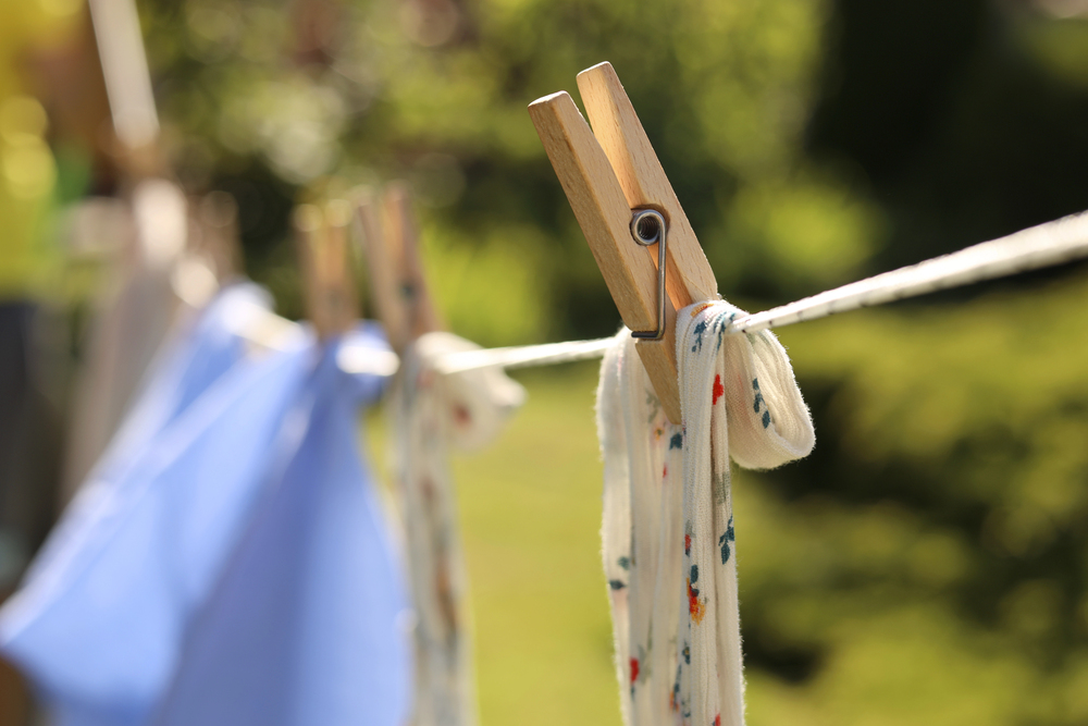 Close-up of a wooden clothes peg securing a white fabric with small colourful patterns to a washing line outdoors, with other laundry items in the blurred background. Bright sunlight and greenery are visible.