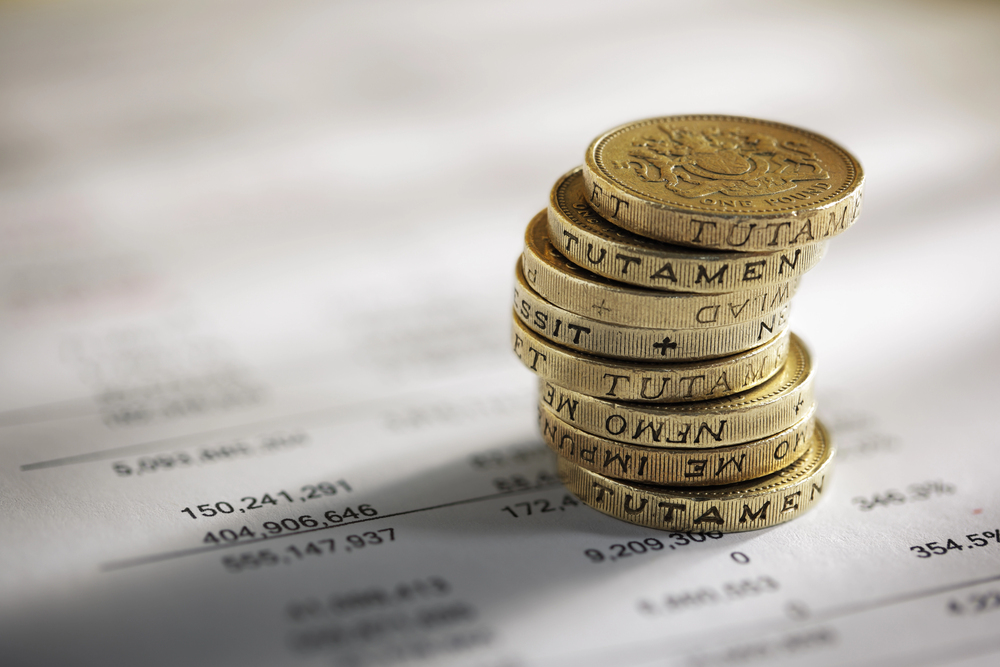 A stack of British one-pound coins is placed on financial documents, with numbers and text visible in the background. The coins are in sharp focus whilst the paperwork is blurred.