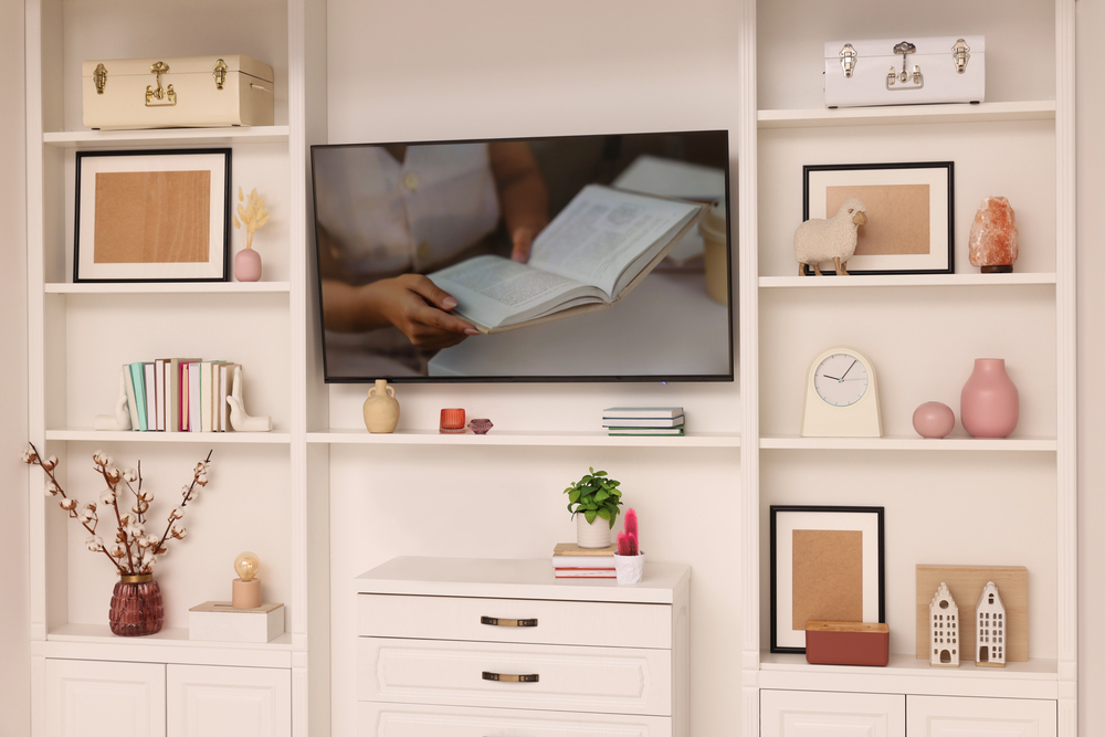 A modern white living room shelf with books, framed art, decorative boxes, and ornaments. A TV in the centre displays a person holding an open book. Pink and beige accents add warmth to the minimalist decor.