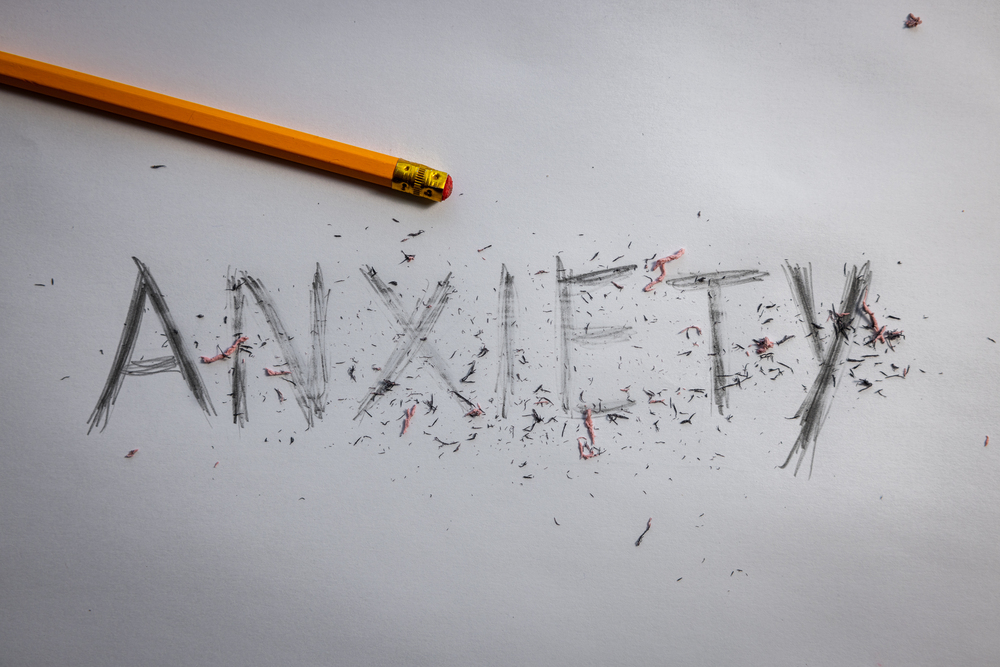 A yellow pencil lies on paper with the word ANXIETY heavily rubbed out but still visible, surrounded by rubber shavings and pencil marks.