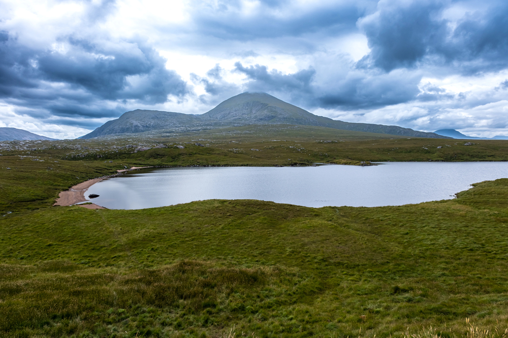 A tranquil lake surrounded by green grassy hills, with a mountain rising in the background under a dramatic cloudy sky. The landscape appears remote and unspoilt, evoking a sense of solitude and peace.