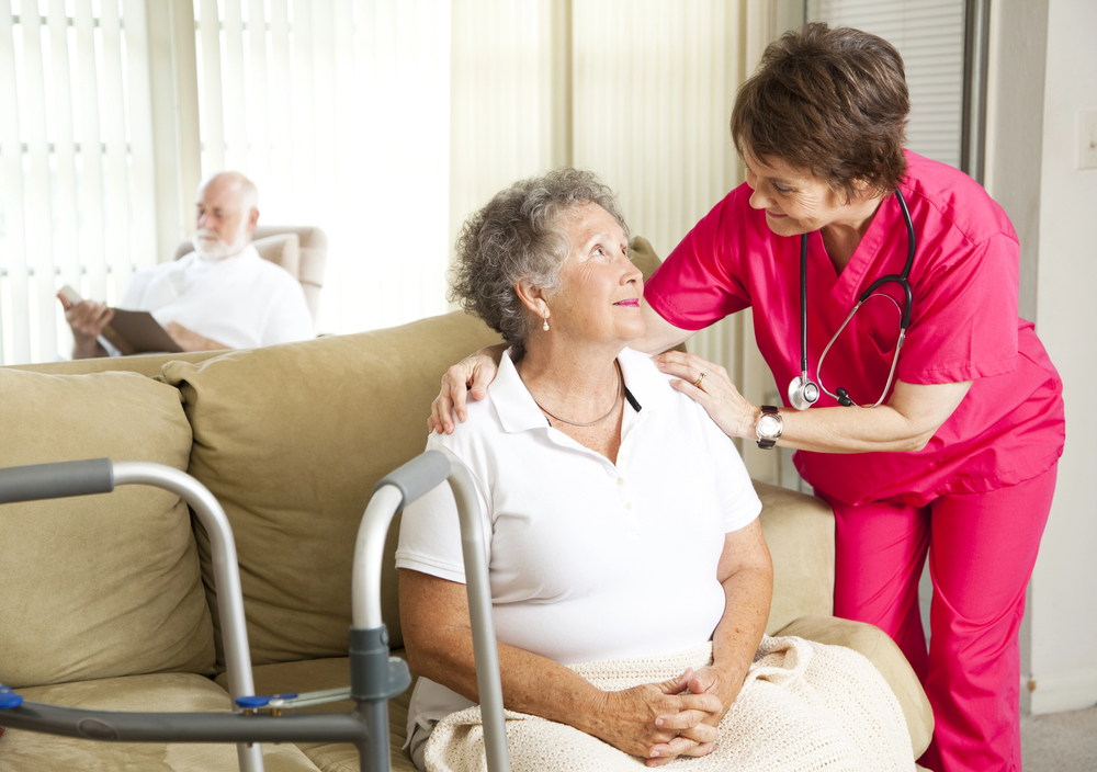 A nurse in pink scrubs smiles and talks to an elderly woman sitting on a sofa with a walking frame nearby. An older man sits in the background reading, in a bright, comfortable living room.