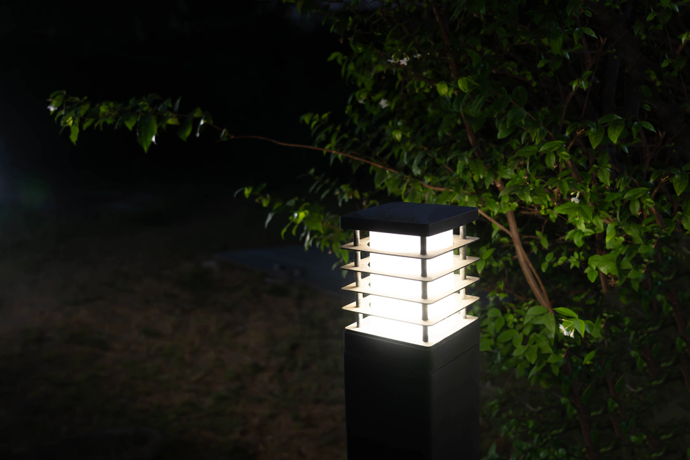 A modern outdoor lamp post glows brightly at night, illuminating nearby green leaves and casting light on the surrounding dark garden.