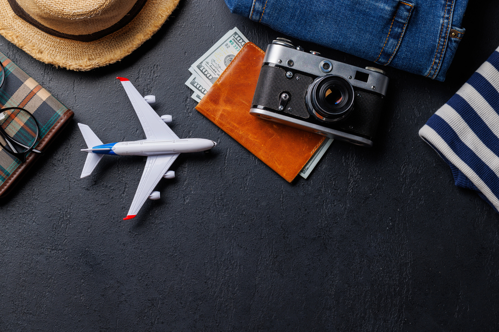 Flat lay of travel essentials including a toy aeroplane, camera, passport, cash, glasses, hat, checked notebook, jeans, and striped jumper on a dark surface.