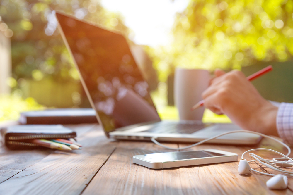 A person works on a laptop at an outdoor wooden table, holding a pen and a coffee mug. Nearby are coloured pencils, a notebook, a mobile phone, and earphones, with greenery and sunlight in the background.