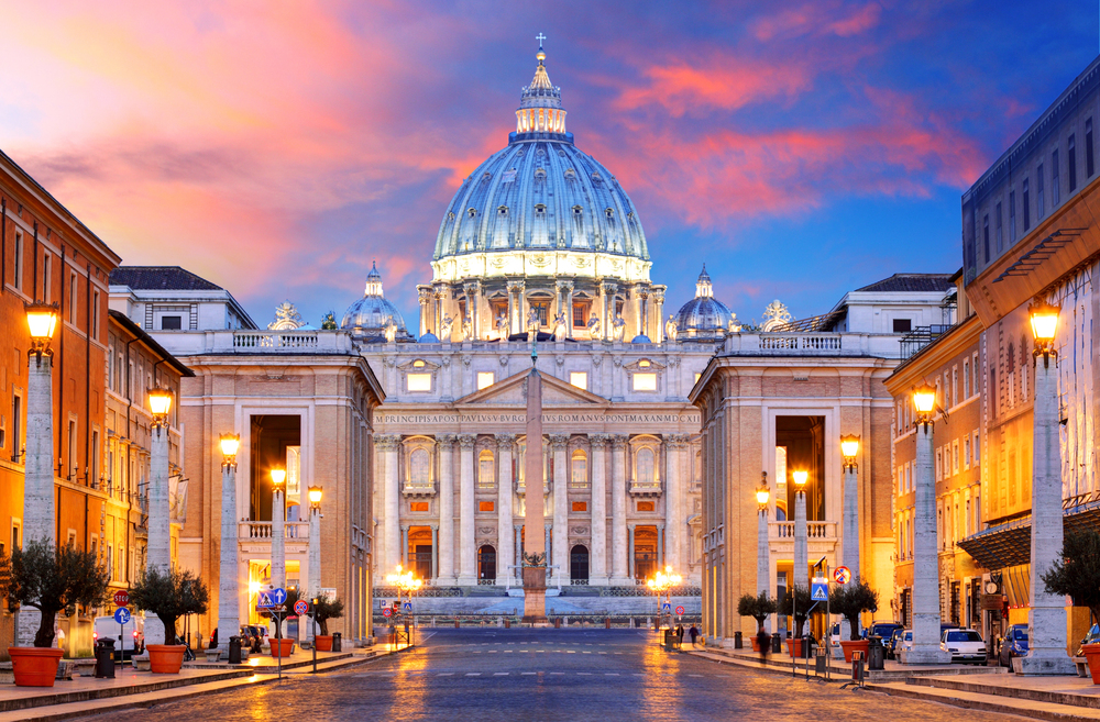 St. Peter’s Basilica in Vatican City is illuminated at dusk, with its grand dome and façade glowing under a vibrant pink and blue sunset sky. Street lamps line the symmetrical road leading to the basilica.