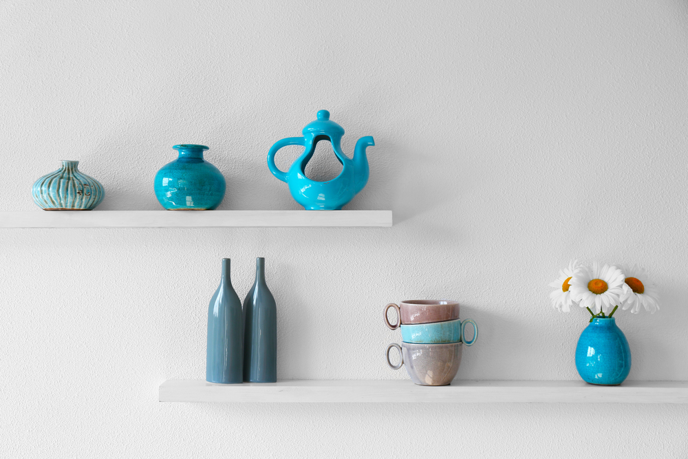 Two white wall shelves display blue ceramic vases, a teapot, two bottles, stacked mugs, and a blue vase with white daisies, all set against a plain white background.