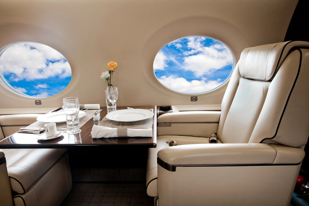Luxurious private jet interior with cream leather seats, a dining table set with elegant crockery and glassware, and two large windows showing a blue sky with clouds. A single rose in a vase decorates the table.