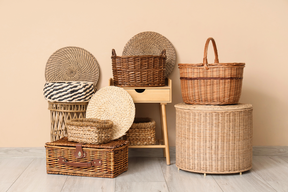 Several woven baskets of different shapes and sizes are stacked and arranged on a wooden stool and wicker furniture against a beige wall on a light wooden floor.