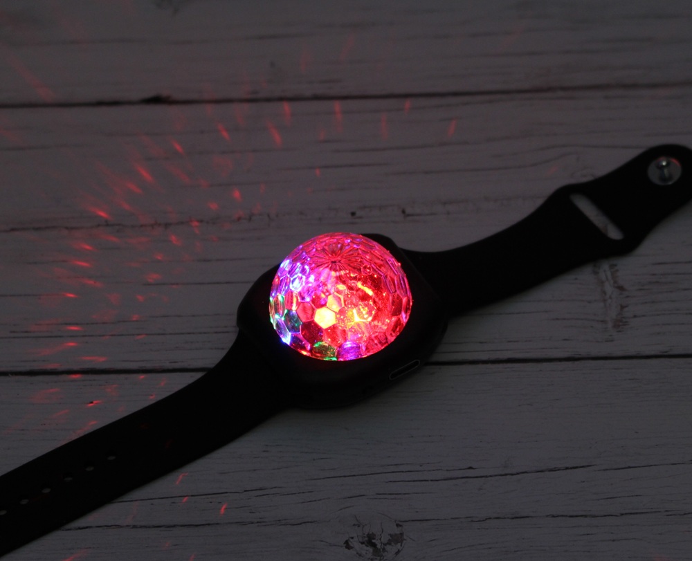 A black wristband with a dome-shaped, faceted light that glows red and multicoloured, casting vibrant patterns on a white wooden surface.
