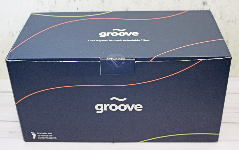 A dark blue box with the word “groove” in white text on the front and top, containing the original Groove adjustable pillow. The box features curved coloured lines and a small carbon footprint icon in the lower left corner.