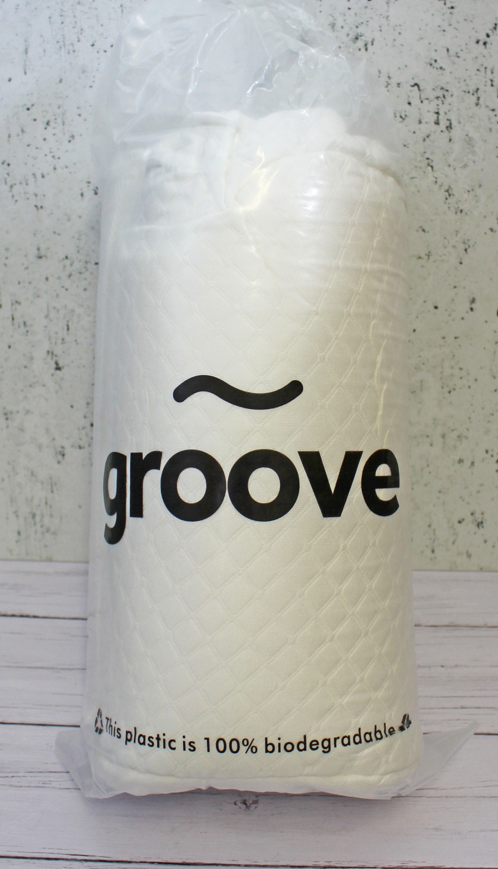 A white quilted fabric roll in a clear plastic bag labelled groove. The bag states, This plastic is 100% biodegradable. The background is a light, textured wall and wooden surface.