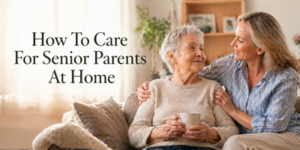 An older woman holding a mug sits on a sofa while a younger woman hugs her and smiles. Text on the image reads: How To Care For Elderly Parents At Home. They appear happy and comfortable in a cosy living room.