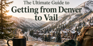A steaming mug, notebook, gloves, and a rucksack rest on a wooden table overlooking a snowy mountain town with a winding road. Text reads: The Ultimate Guide to Getting from Denver to Vail.