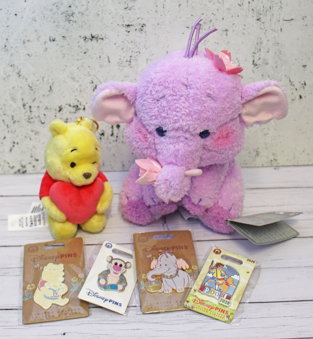 A small Winnie the Pooh soft toy with a red heart and a larger purple Heffalump soft toy are displayed behind four Disney character enamel badges—favourites that inspire repeat purchases—on a white wooden surface with a textured white background.