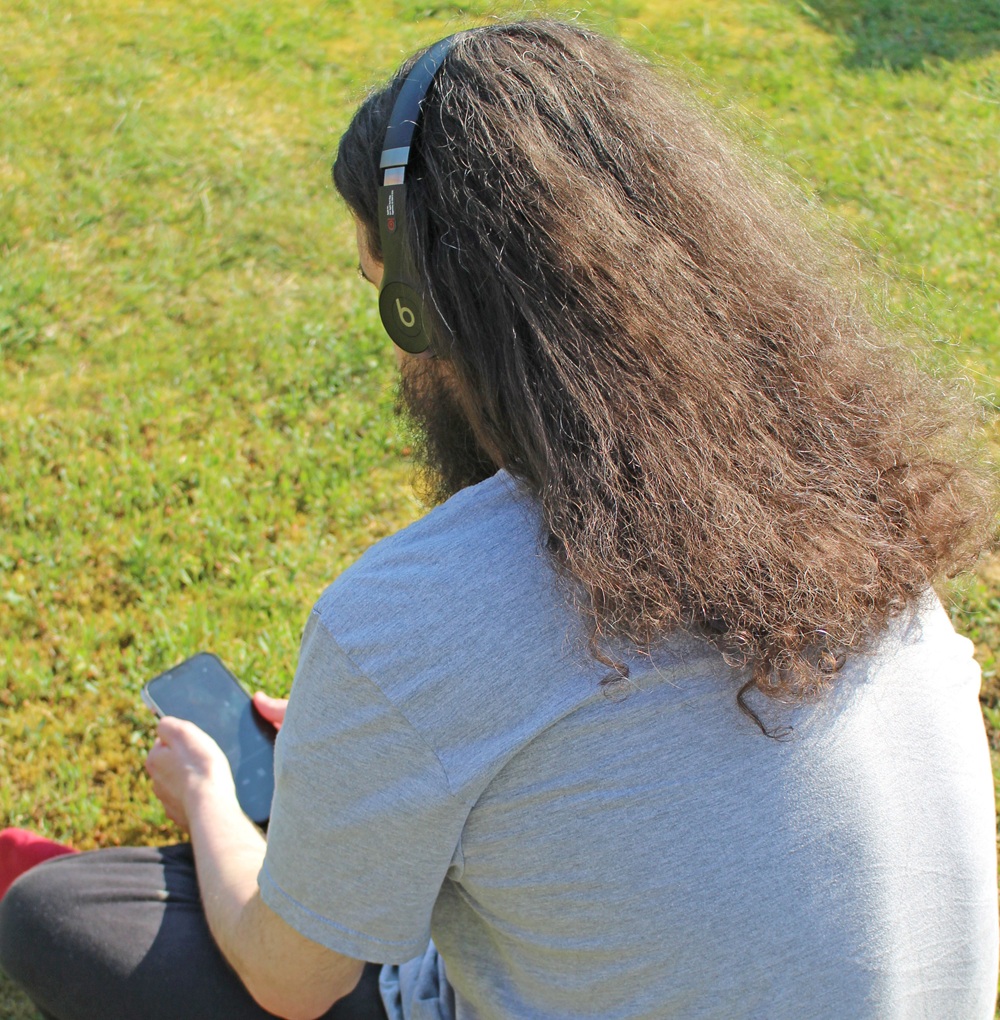 A person with long brown hair and a grey T-shirt sits on grass, listening to music with black Beats headphones—an ideal scene for Father's Day Gift Ideas—while holding a mobile and facing away from the camera.
