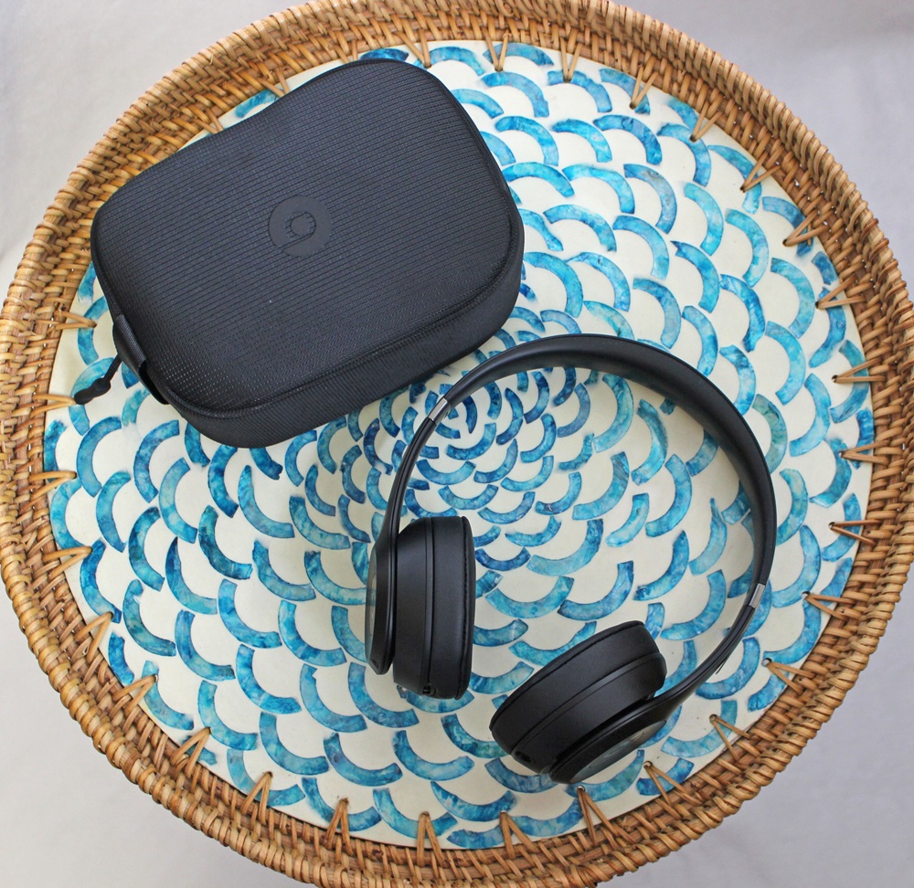 A pair of black over-ear headphones and a matching black carrying case rest on a round, woven rattan tray with a blue and white patterned base.