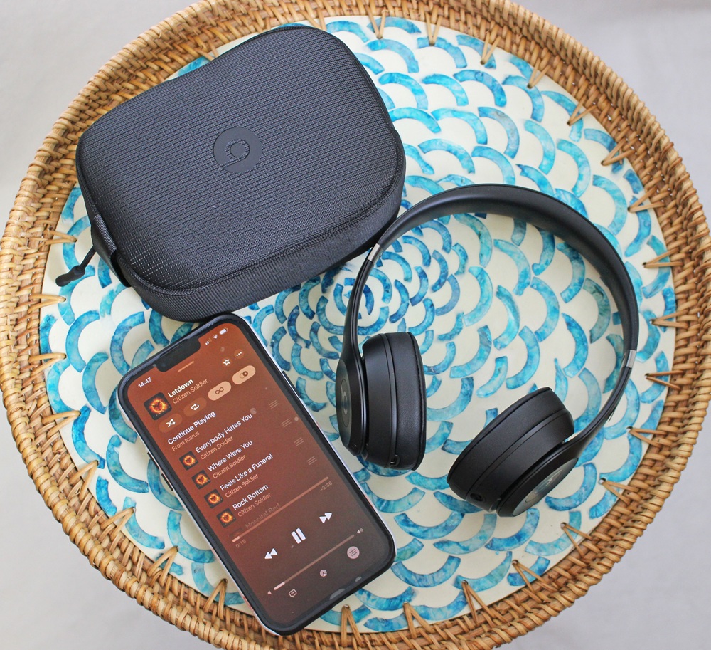 A smartphone with a music playlist on screen, black wireless headphones, and a black carrying case rest on a round woven table with a blue and white patterned top.