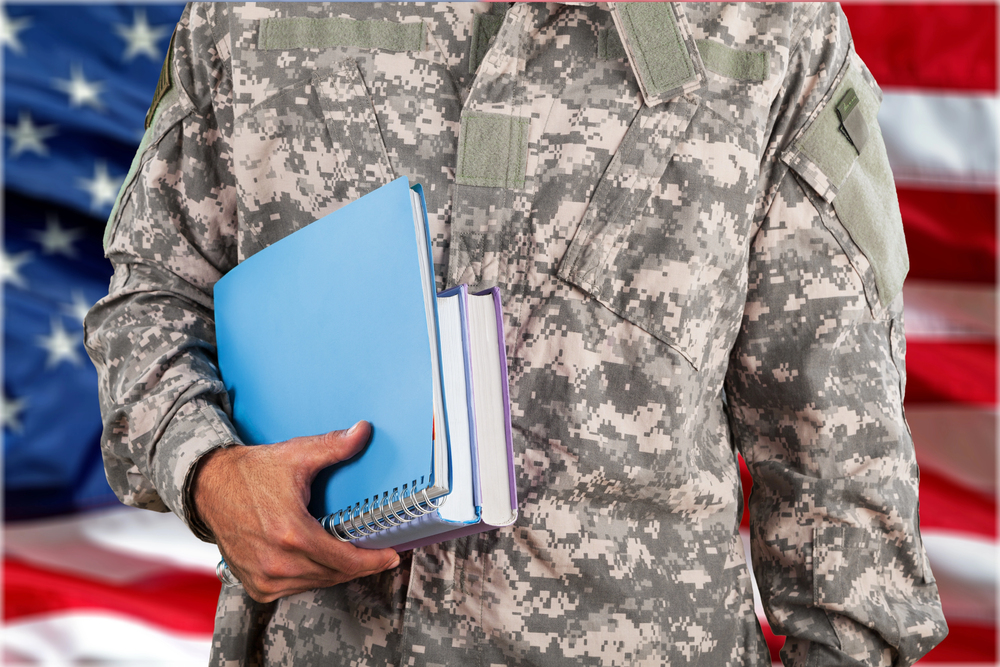 A person in military camouflage uniform holds books and a notebook in front of a blurred American flag background.