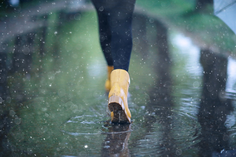 Person wearing yellow wellington boots walking through a puddle on a rainy day, with water splashing and raindrops visible in the air. The background is blurred with green and grey tones.