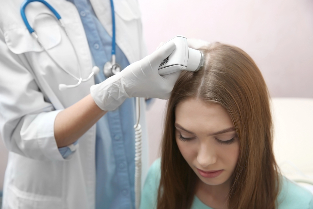 A doctor wearing gloves uses a handheld device to examine the scalp of a young woman with long brown hair. The woman looks downwards while the doctor conducts the examination.