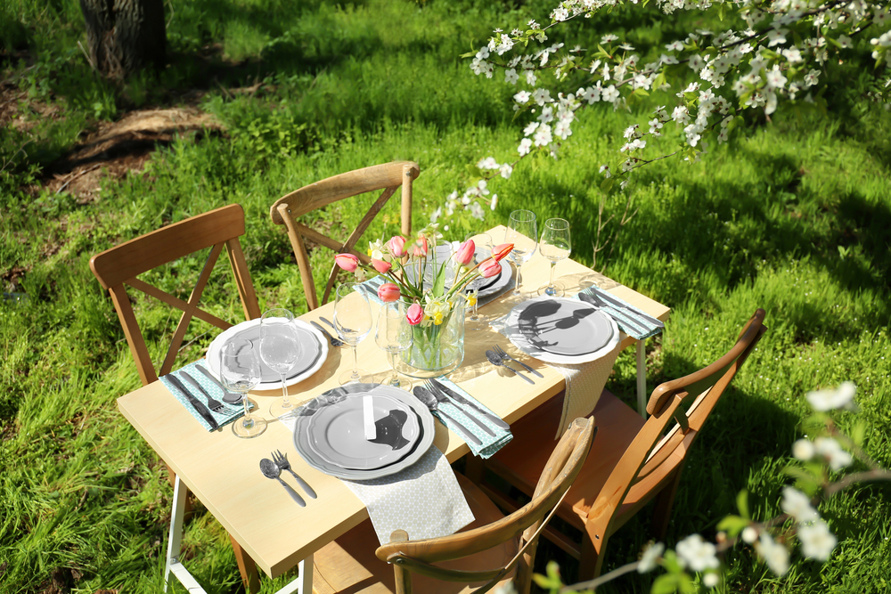 A small wooden table with four chairs is set outdoors on green grass. The table is decorated with a vase of pink tulips, plates, glasses, cutlery, and serviettes under blossoming tree branches.