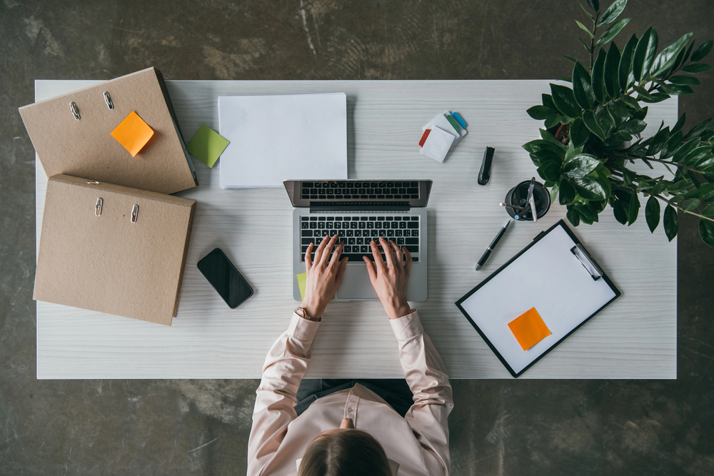 Top view of a person typing on a laptop at a tidy white desk with folders, a mobile phone, clipboard, pens, sticky notes, papers, and a potted plant.