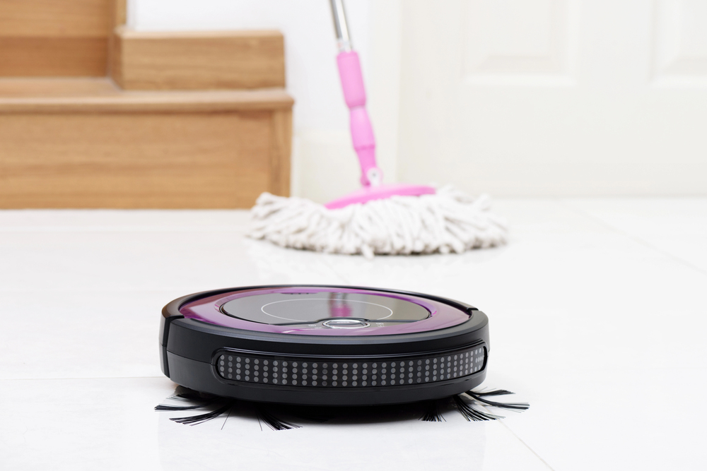 A robotic vacuum cleaner moves across a white tiled floor near stairs, with a mop and a door in the background.