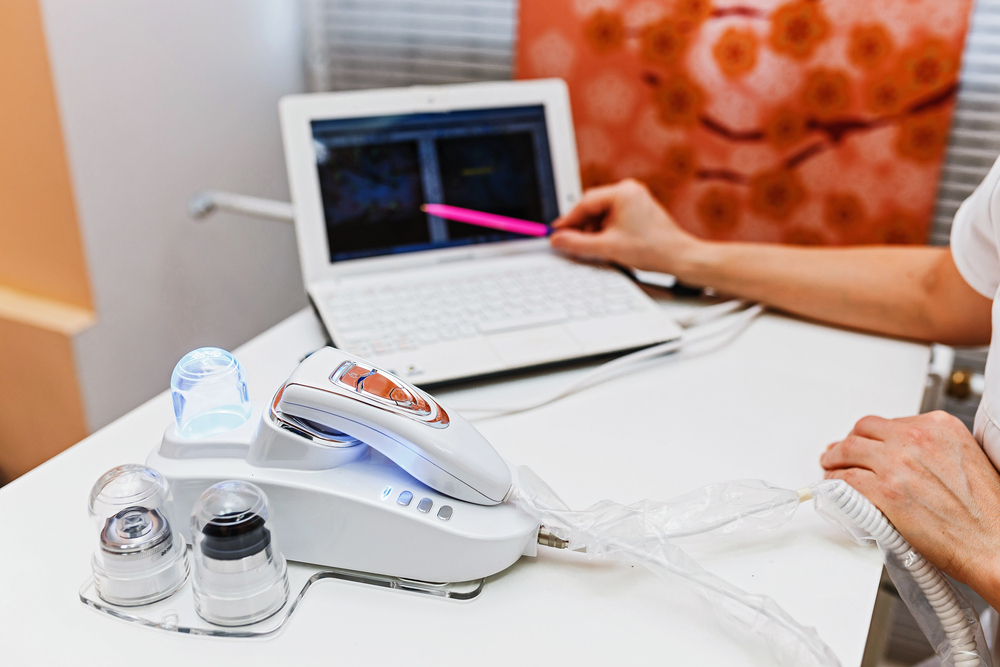 A person uses a laptop with one hand while the other hand rests on a cosmetic skincare device with multiple attachments, placed on a white desk. The background includes a patterned orange and white cushion.