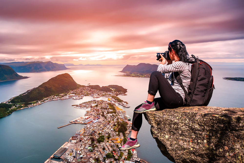 A woman with a rucksack sits on a cliff edge photographing a coastal town below at sunset, with mountains and water in the background.