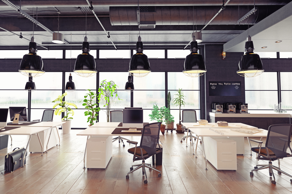 A modern, open-plan office with white desks, black chairs, large hanging lamps, indoor plants, and floor-to-ceiling windows letting in natural light. A coffee station is visible in the background.