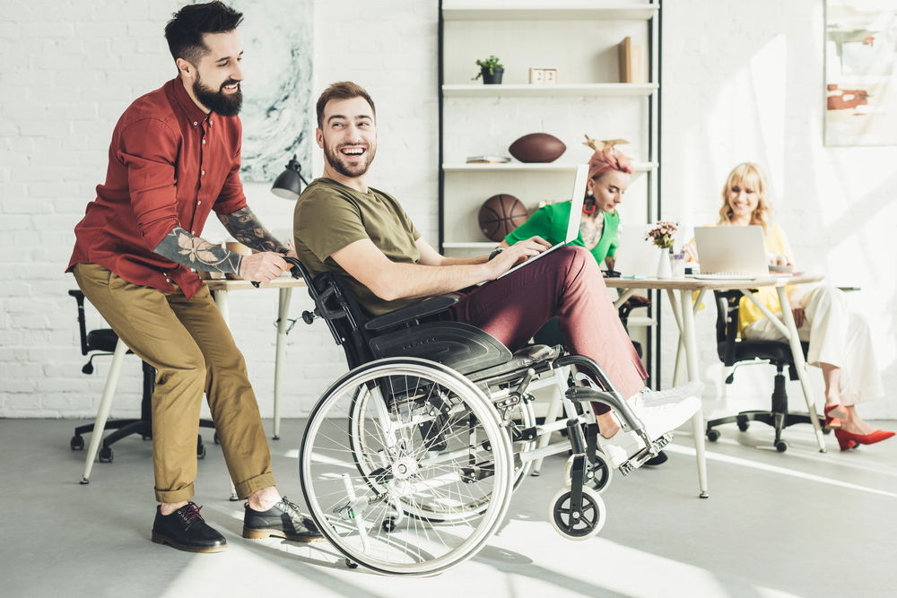 A smiling man in a wheelchair is pushed by a cheerful colleague in a bright office where two women are working at desks in the background.