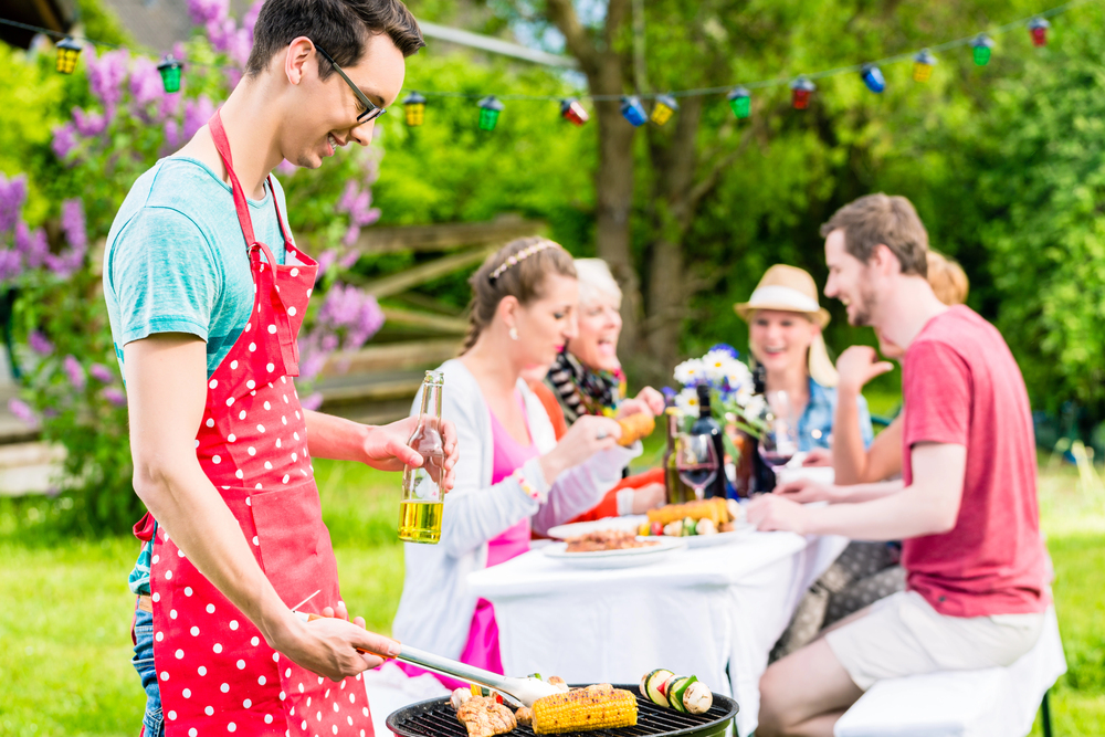 A person wearing a red polka-dot apron grills food outdoors while holding a drink. In the background, four people sit at a picnic table, enjoying food and talking in a garden setting with festive lights.
