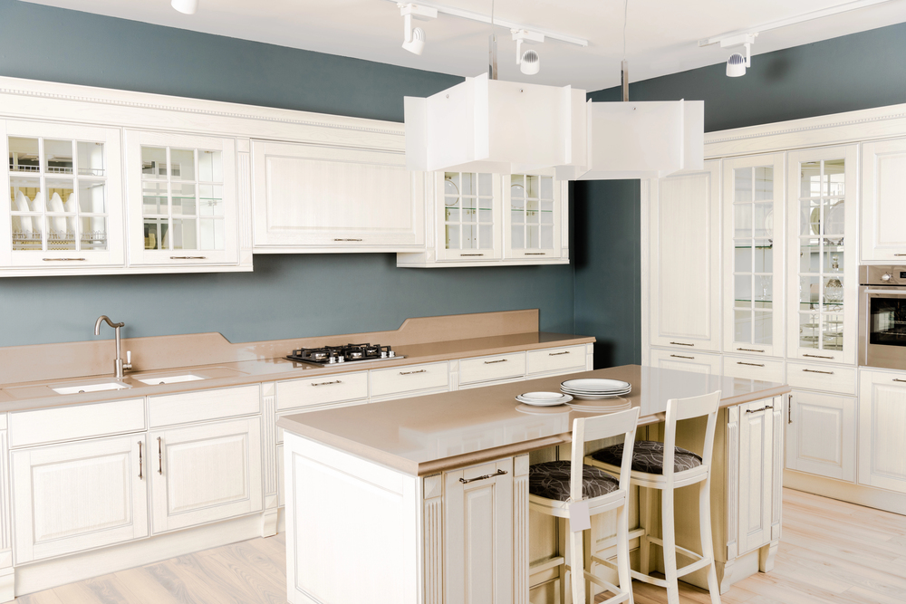 A modern kitchen with white cupboards, beige worktops, an island with two chairs, a built-in hob, glass-fronted cupboards, and pendant lights hanging above the island.