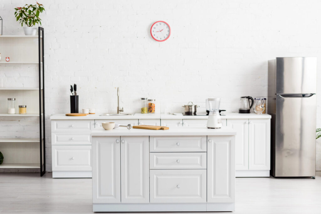 A modern kitchen with white cupboards, a kitchen island, stainless steel fridge, and various appliances on the worktop. A clock hangs on the white brick wall above the sink, and shelves hold jars and kitchenware.