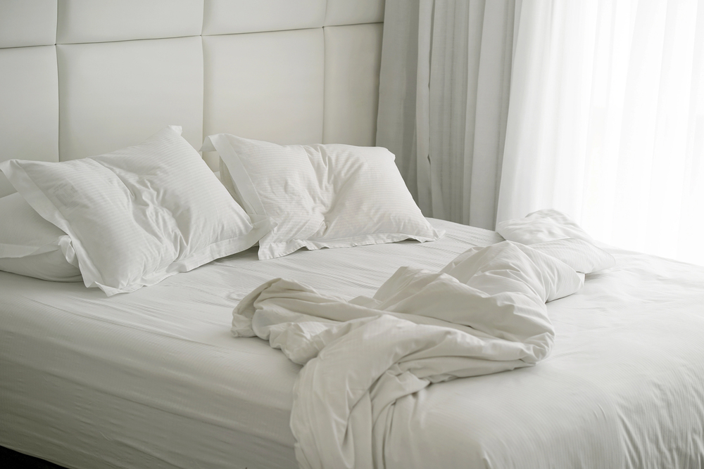 A bed with white sheets, two pillows, and a blanket that appears unmade. The headboard is upholstered and white, and sheer curtains let in soft natural light from a nearby window.