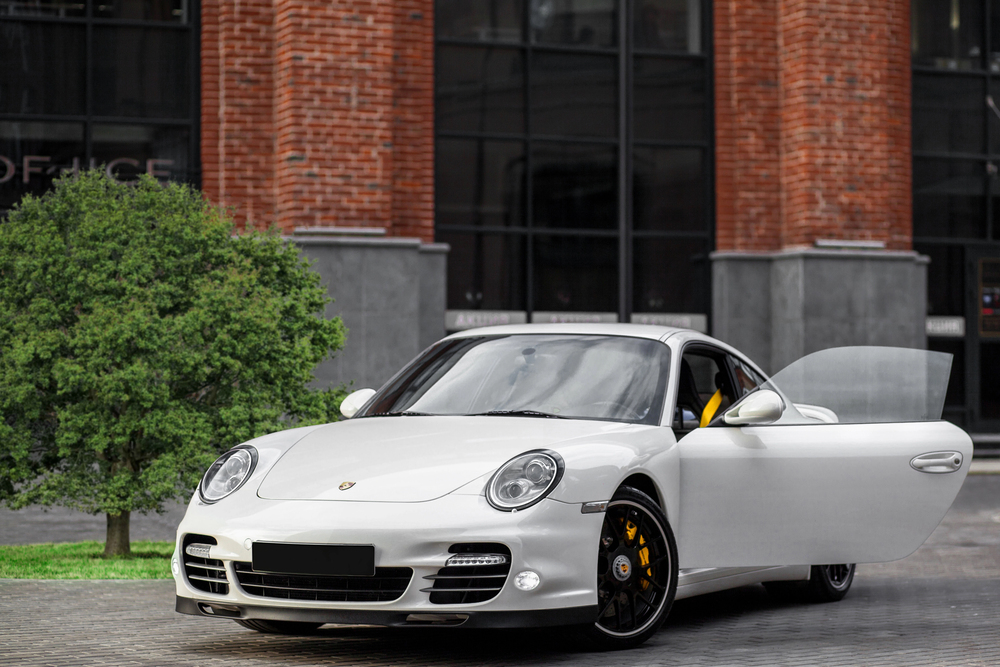 A silver Porsche sports car with its driver’s side door open is parked on a cobbled street in front of a red brick building and a small green tree.