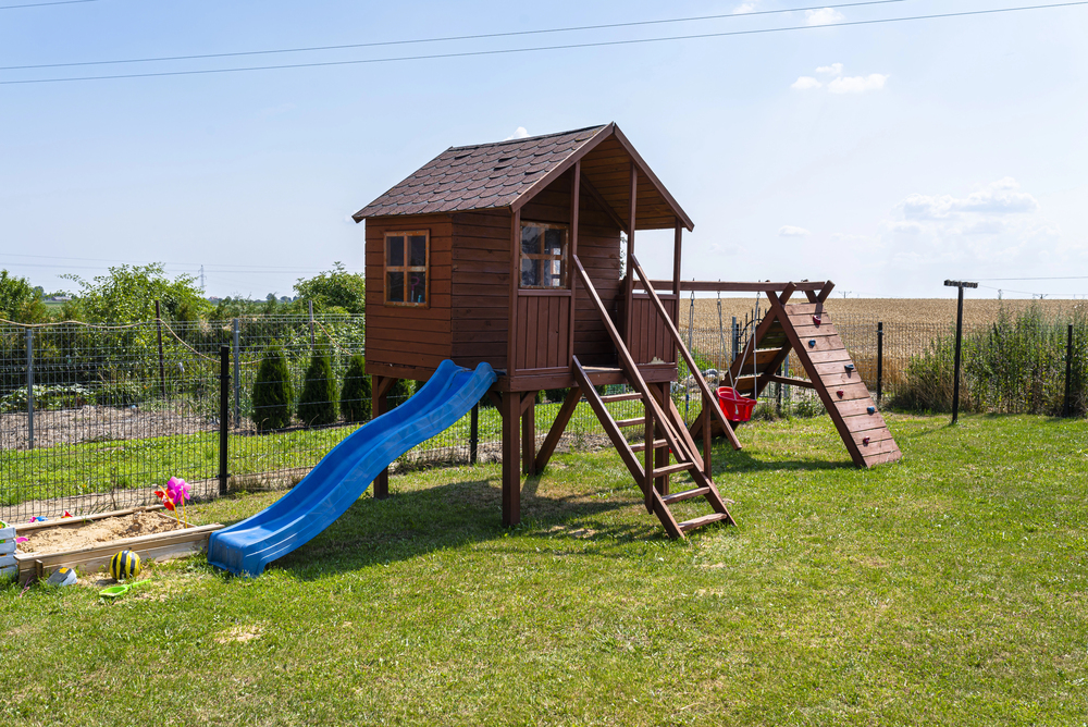 A wooden playhouse with a slide, ladder, climbing wall, and sandpit stands on a grassy lawn, surrounded by a wire fence and open fields under a partly cloudy sky.