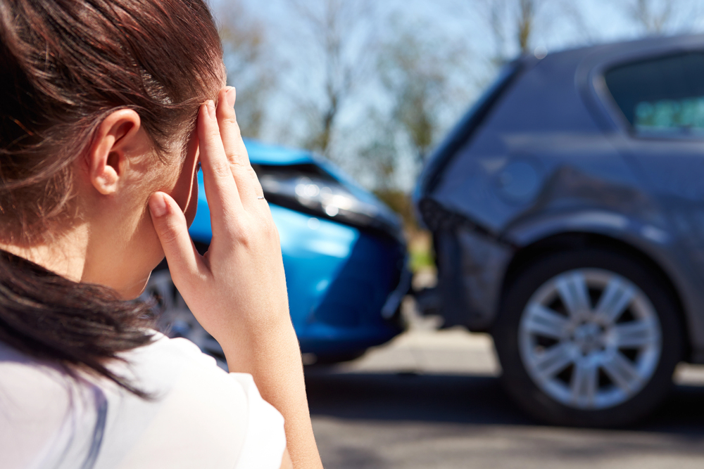 A person holds their head in distress while looking at a rear-end car accident involving a blue car and a grey car on a sunny day.