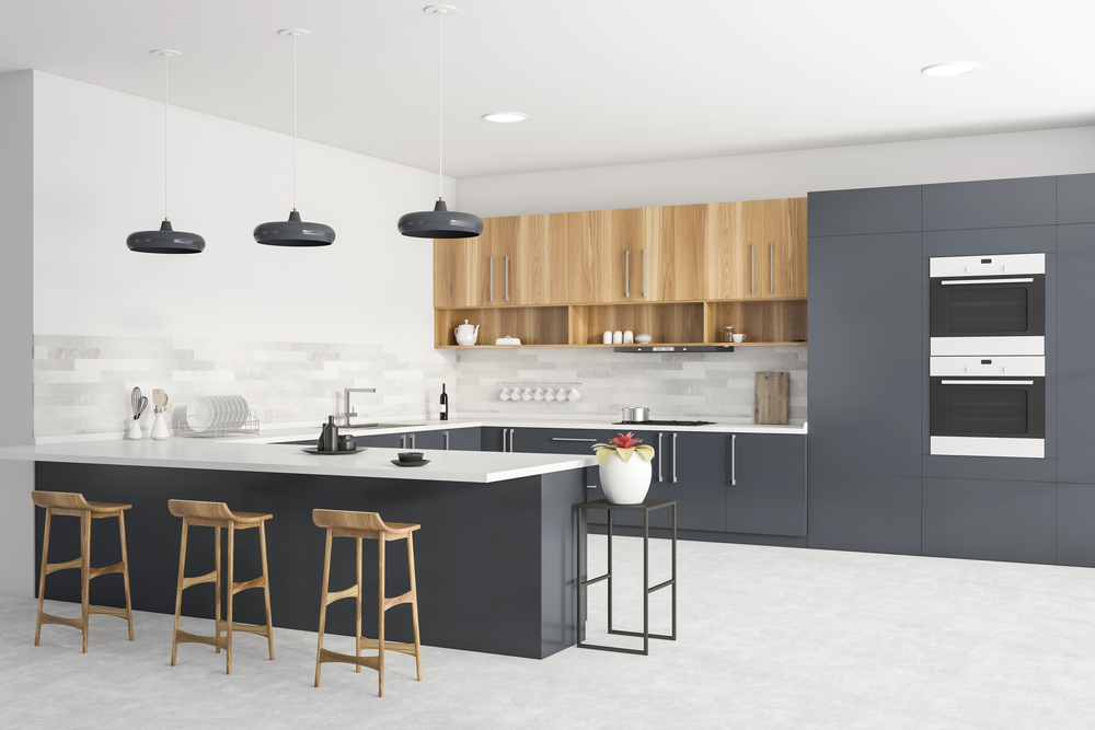 Modern kitchen with dark lower cabinets, light wood upper cabinets, a white island with three wooden stools, black pendant lights, built-in ovens, and minimalist decor including a plant and crockery.