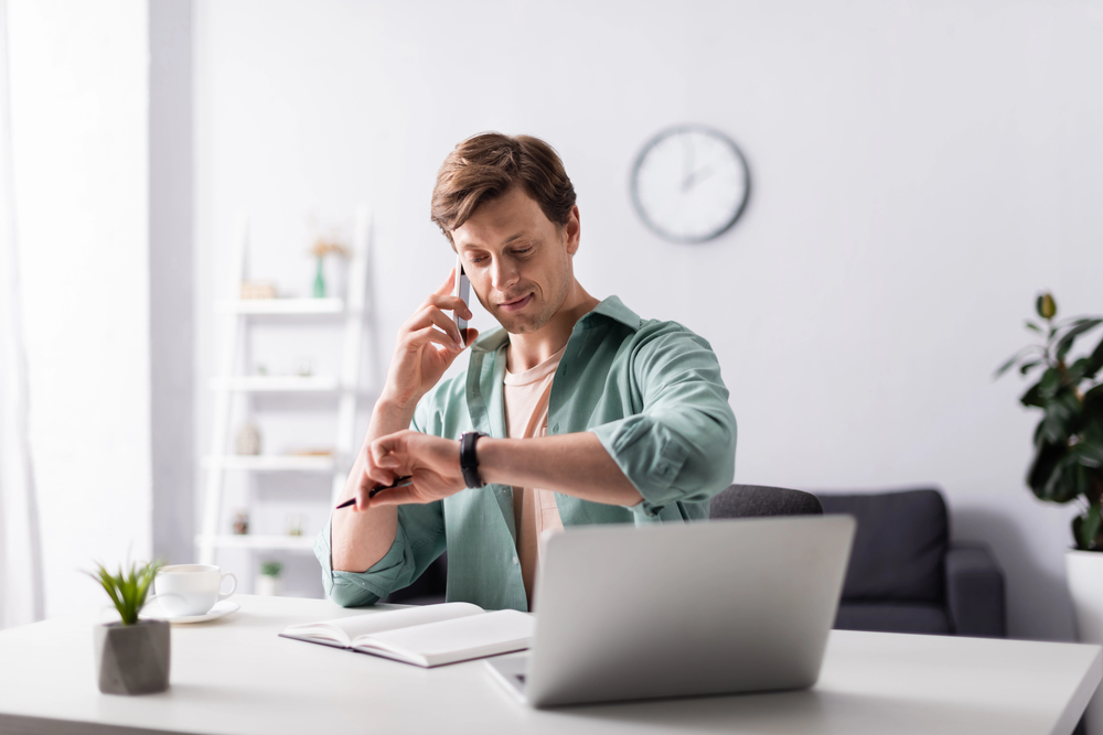 A man sitting at a desk with a laptop, talking on the phone whilst checking his watch. There is an open notebook on the desk, and the background features a clock, shelves, and a plant.
