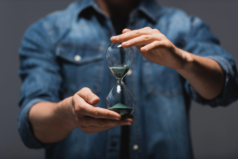 A person in a denim shirt holds an hourglass with green sand, with one hand supporting the base and the other adjusting the top, against a neutral background.