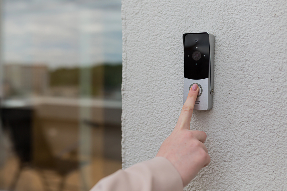 A person presses the button on a modern video doorbell mounted on a white textured wall, with a blurred outdoor background visible.
