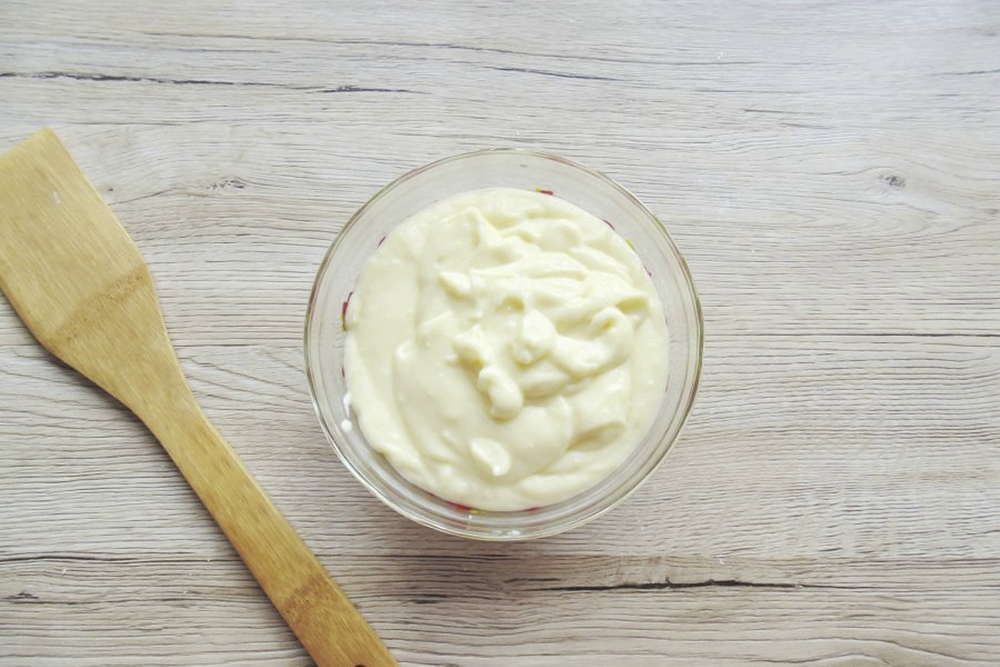 A glass bowl filled with creamy white sauce or batter sits on a light wooden surface next to a wooden spatula.