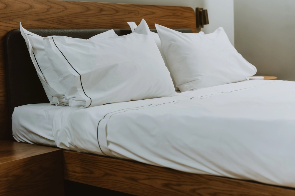 A neatly made bed with white sheets and two pillows featuring black piping, set against a wooden headboard and accompanied by a wooden bedside table.