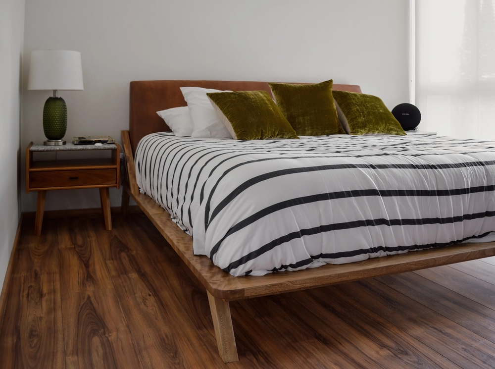 A modern bedroom with a wooden bed frame, striped black and white bedding, green velvet cushions, a wooden bedside table with a lamp, and a smooth wooden floor. Light streams in from a window on the right.